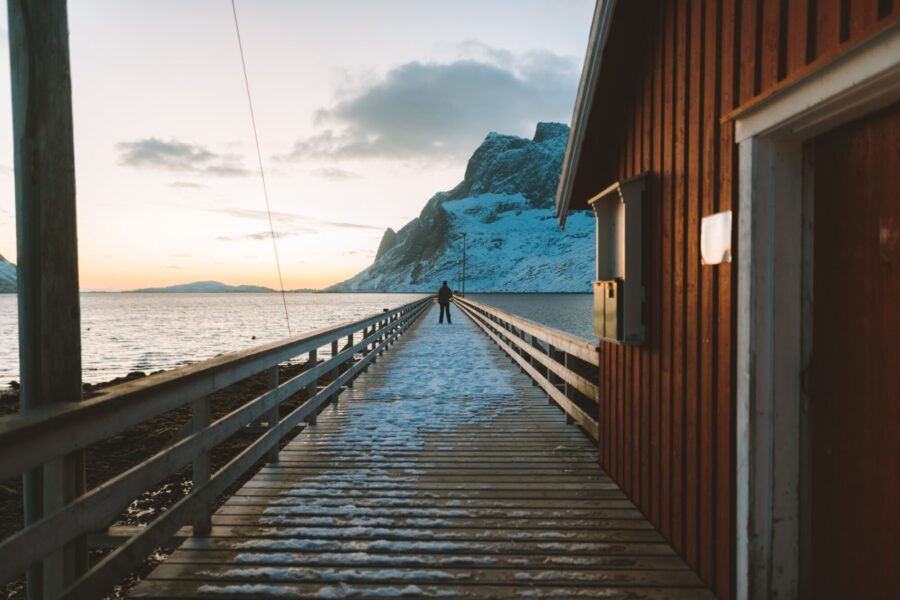 En brygge ved siden a et rødt hus, hvor en person står midt på bryggen i det fjerne