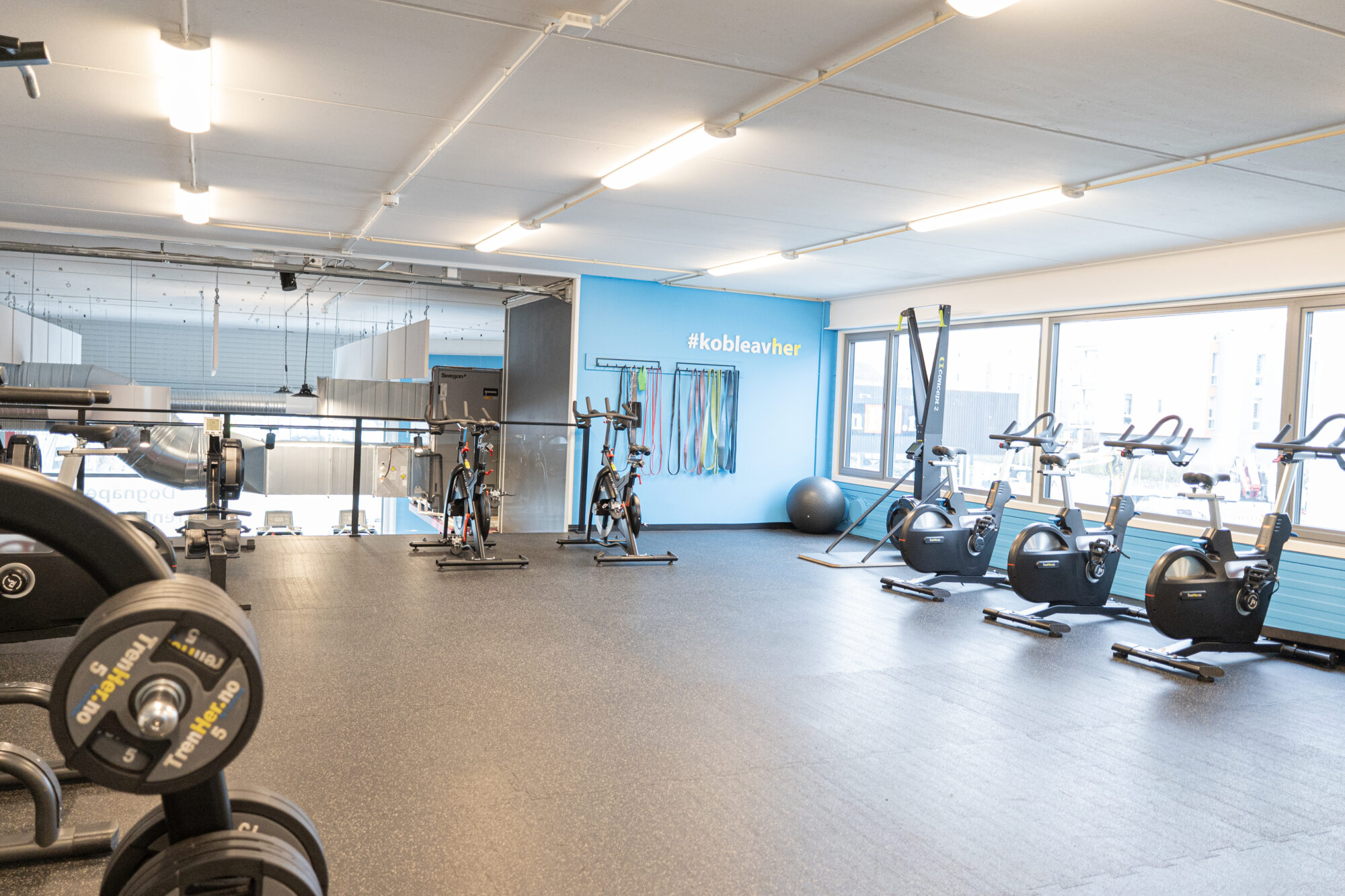 Lyst treningsområde i øverste etasje hos TrenHer Nardo, med spinningsykler, romaskin, SkiErg og strikk for funksjonell trening. Blå vegg med teksten #kobleavher.