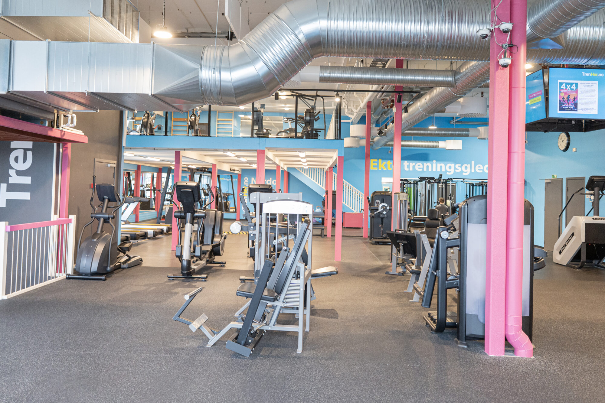 Oversiktsbilde av hovedsalen på TrenHer Nardo, med apparater, ellipsemaskiner og synlig mesanin med ekstra treningsutstyr. Senterets slagord “Ekte treningsglede” på blå vegg.