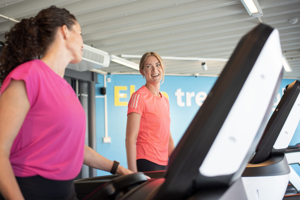 To kvinner trener sammen på tredemølle og smiler til hverandre i et lyst og moderne treningssenter.